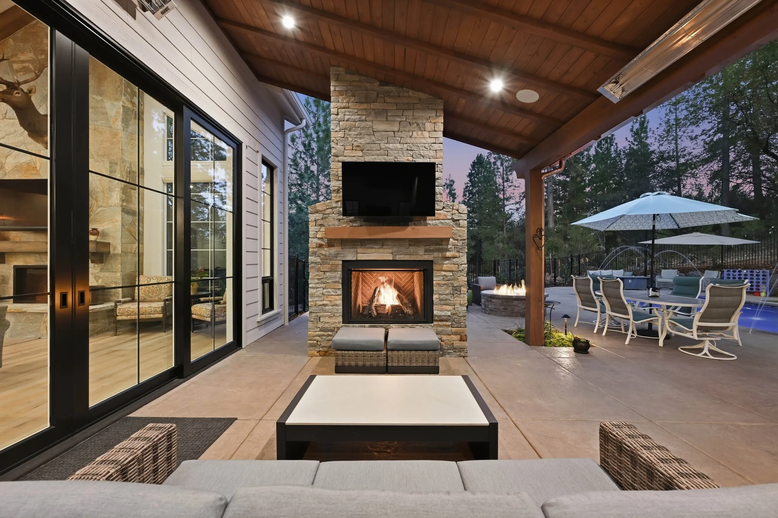 Covered patio with stone outdoor fireplace and stained wood ceiling at twilight
